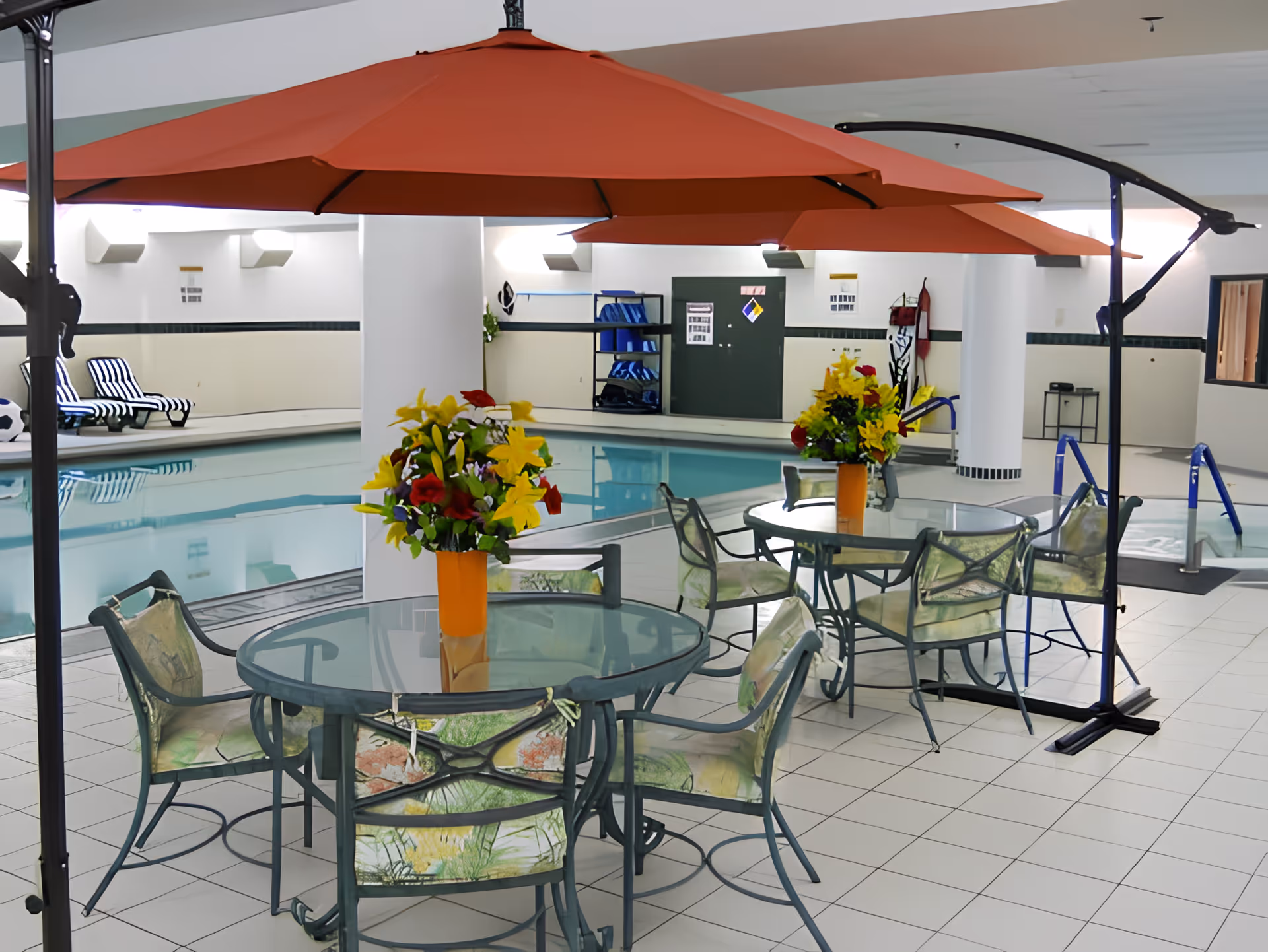 Indoor pool area with two round glass tables, each surrounded by four metal chairs with floral cushions. Each table has a large orange umbrella and a vase of colorful flowers. The pool is visible in the background with striped lounge chairs along the side and pool safety equipment on the walls.