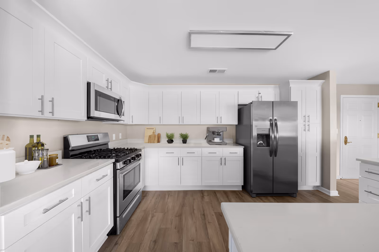 Modern kitchen with white cabinets, stainless steel appliances including a refrigerator, stove, and microwave, wooden flooring, and a white countertop with small decorative plants and kitchen items.