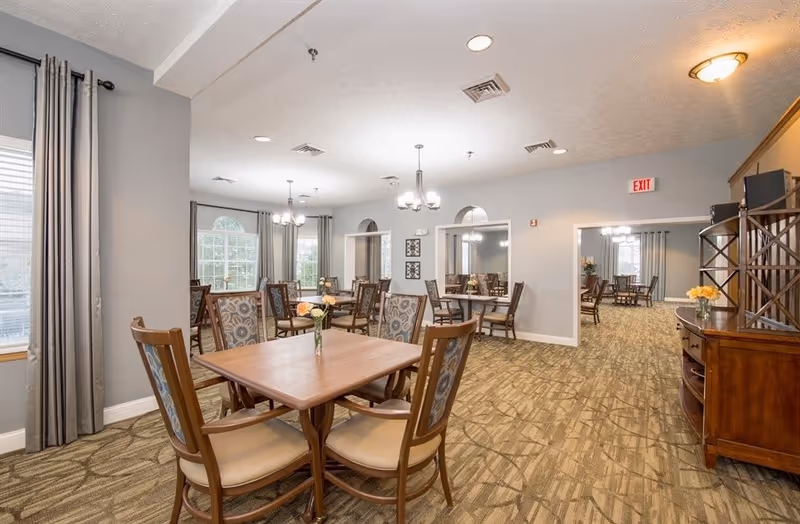 A spacious dining room with multiple wooden tables and chairs arranged neatly. The room has large windows with gray curtains allowing natural light to enter. There are chandeliers hanging from the ceiling and a wooden cabinet with decorative items on the right side. The carpet has a patterned design and the walls are painted light gray.