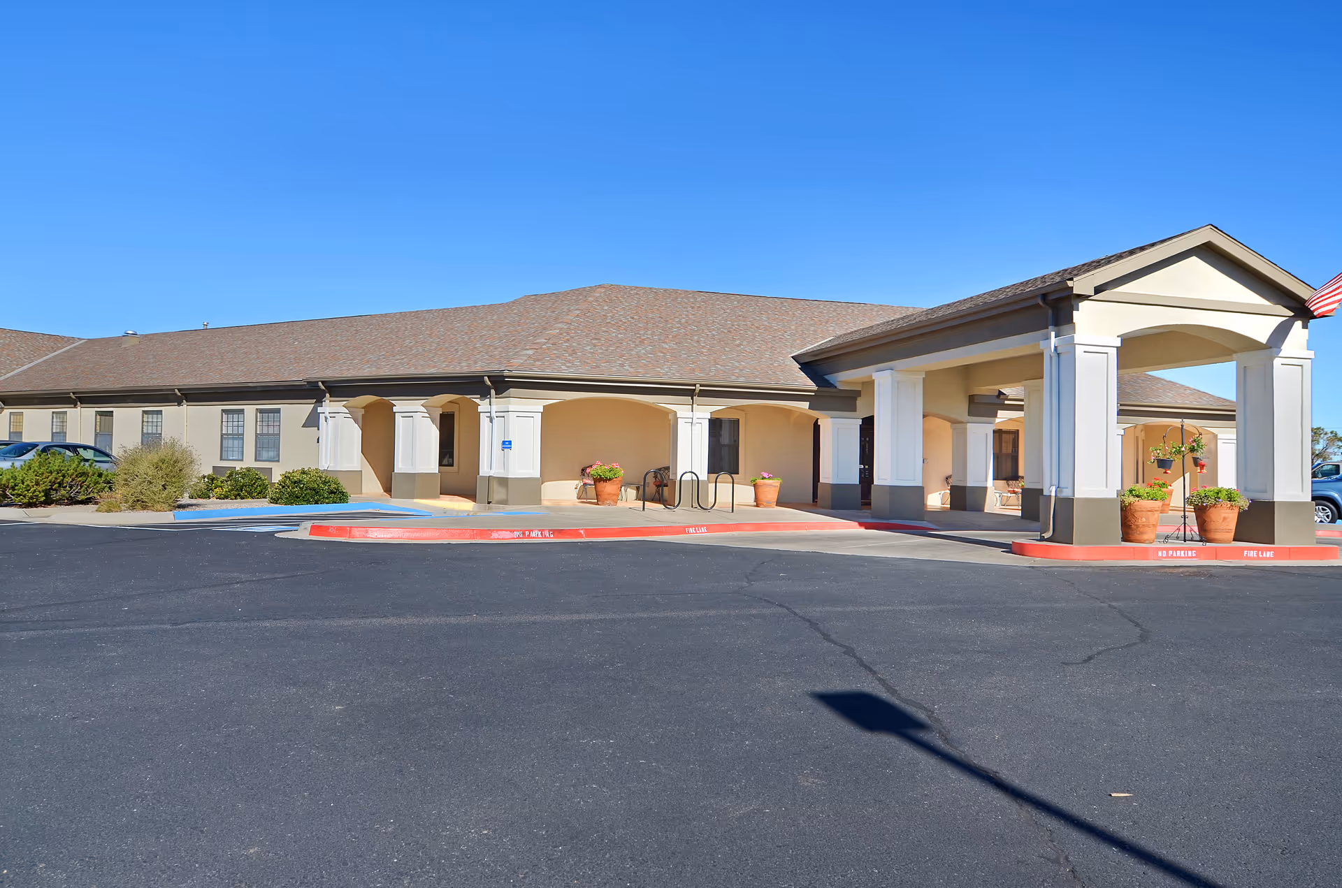 One-story senior living facility front entrance with a covered porte-cochere, potted plants, and a parking/drop-off area under a clear blue sky.