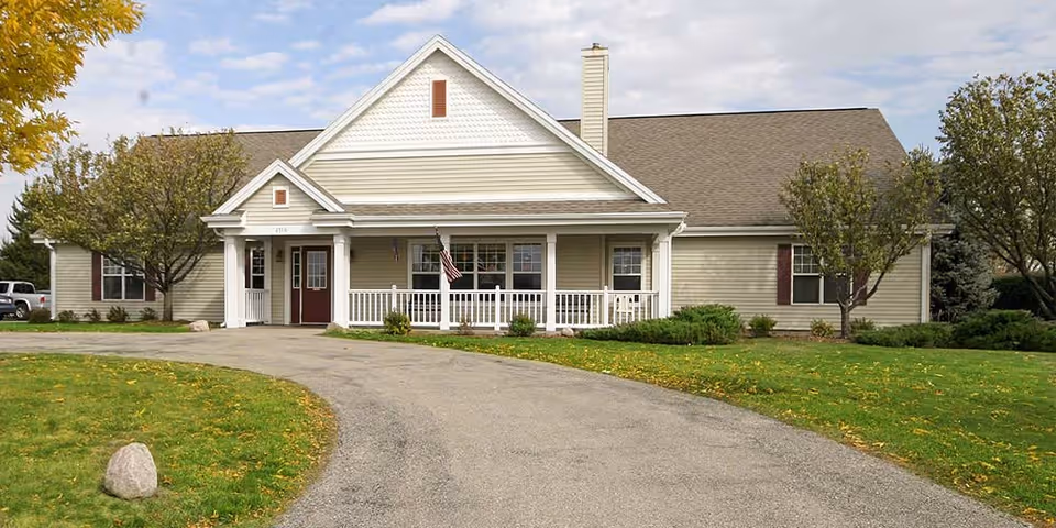 Front exterior view of a single-story senior living facility building with beige siding, a gabled roof, a covered porch with white railings, and an American flag. The driveway curves in front of the building, and there are trees and green grass on either side.