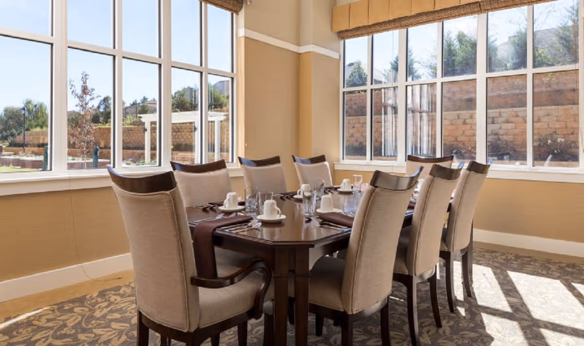 Sunlit dining room with a wooden table set for eight, upholstered chairs, and large windows overlooking an exterior courtyard.
