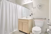 A clean bathroom with a white shower curtain, a wooden vanity with a white sink, a large mirror above the sink, a white toilet with a grab bar next to it, and a wall clock above the toilet.