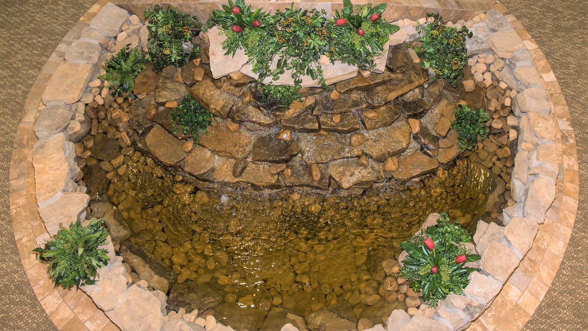Top-down view of an indoor decorative water feature with a stone waterfall surrounded by plants and flowers, set within a circular stone border on a carpeted floor.