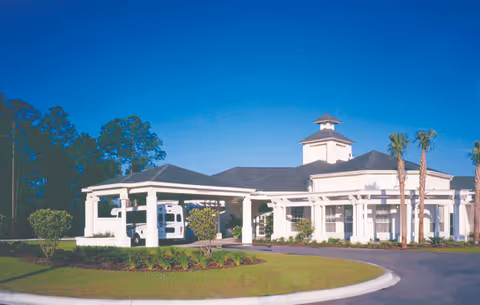 Exterior view of a single-story retirement community building with a covered entrance, white columns, and a small tower on the roof. The building is surrounded by landscaped greenery, including palm trees and bushes, under a clear blue sky.