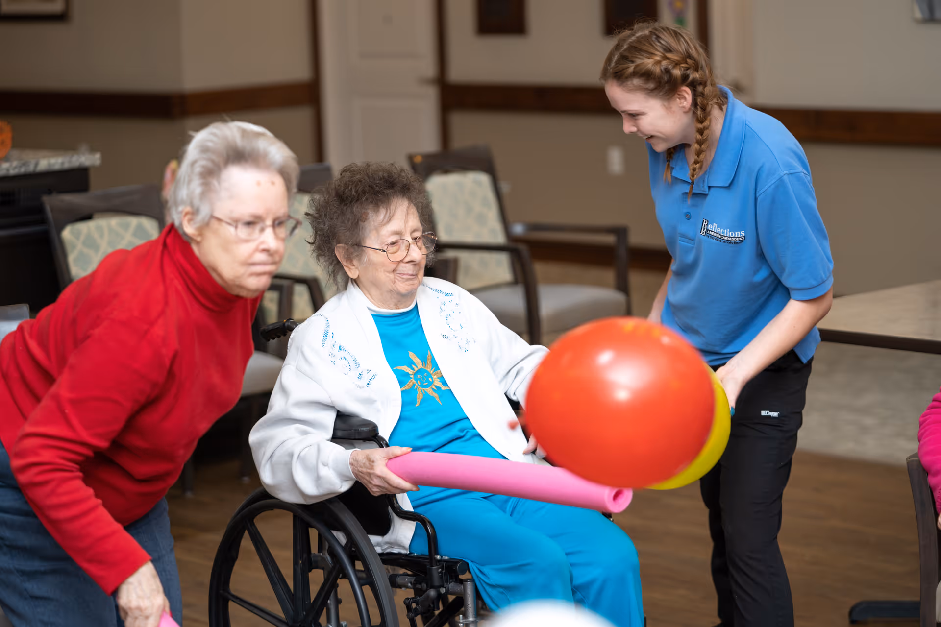 Two elderly women and a young female caregiver are engaged in a playful activity indoors. One elderly woman in a wheelchair holds a pink pool noodle with a large red and yellow ball attached, while the caregiver in a blue shirt smiles and assists. Another elderly woman in a red sweater leans forward, participating in the activity. Chairs and a table are visible in the background.