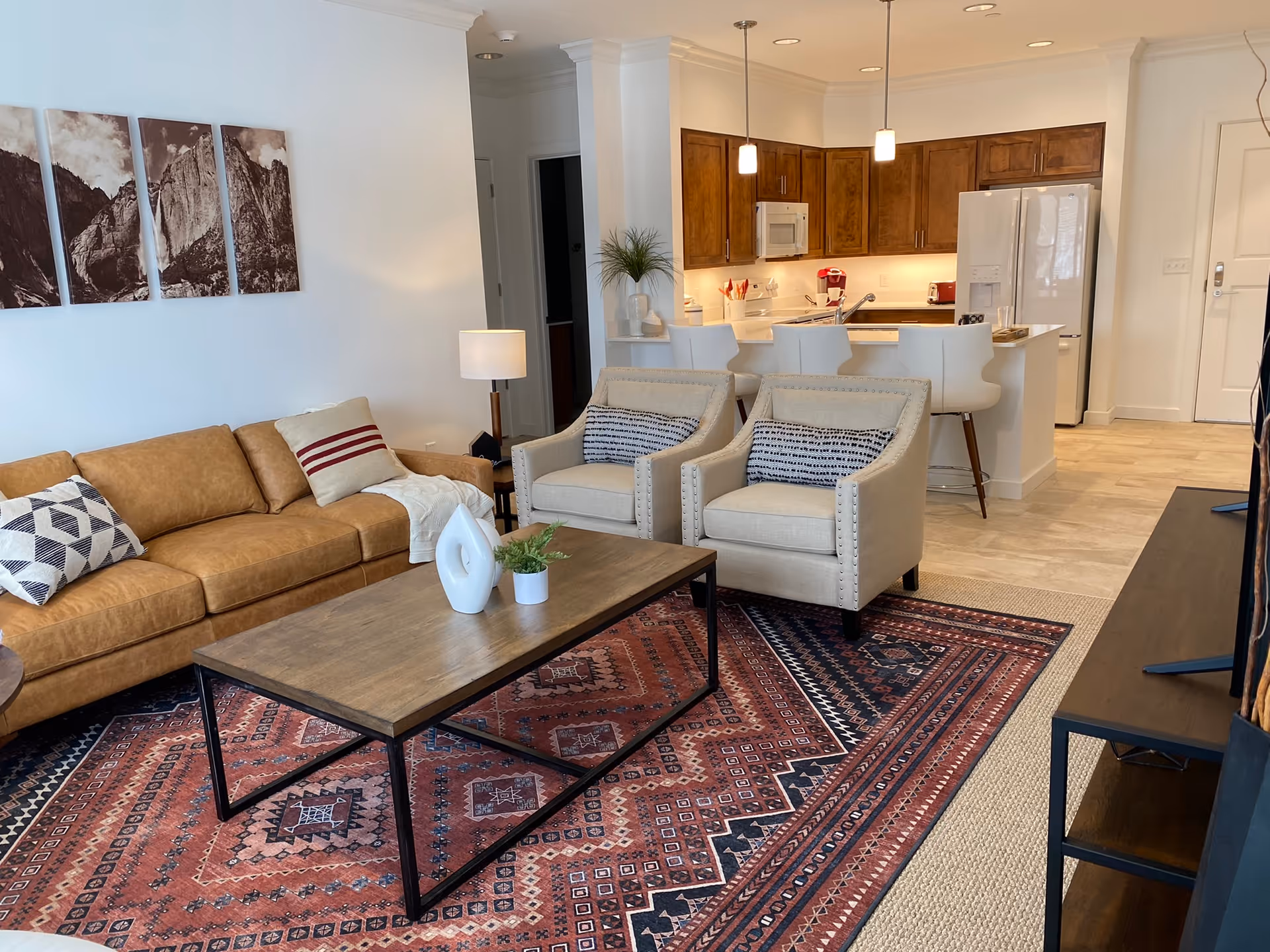 A cozy living room area with a tan leather sofa, two beige armchairs with patterned cushions, and a wooden coffee table on a red patterned rug. The background shows a kitchen with wooden cabinets, white countertops, a refrigerator, and bar stools. There is a floor lamp next to the sofa and a set of four black and white mountain landscape pictures on the wall.