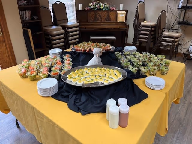 A table covered with a yellow tablecloth and a black cloth centerpiece, displaying trays of deviled eggs and small cups of salad. Plates are stacked on the table, and three bottles of dressing or sauce are placed in front. In the background, there are stacked chairs, a piano, and a bookshelf.