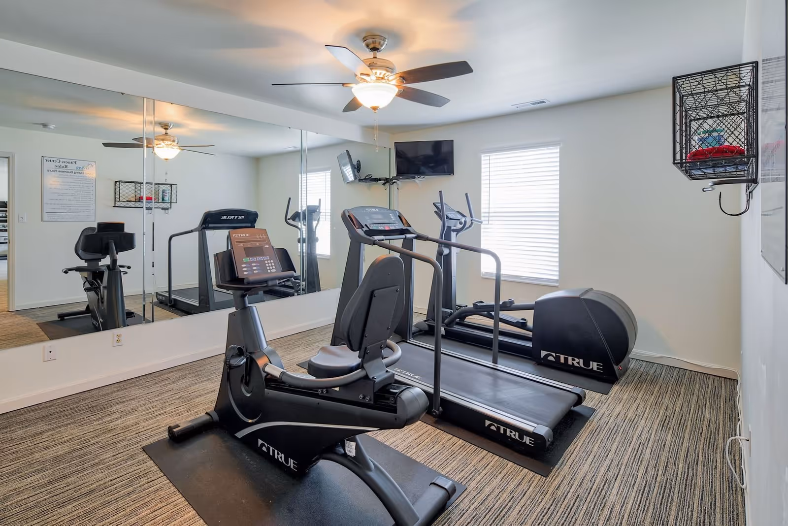 A small fitness center room with exercise equipment including a recumbent bike, treadmill, and elliptical machine. The room has a ceiling fan with light, large mirrors on one wall, a wall-mounted TV, and a window with blinds. There is a wire basket mounted on the wall holding some red towels.