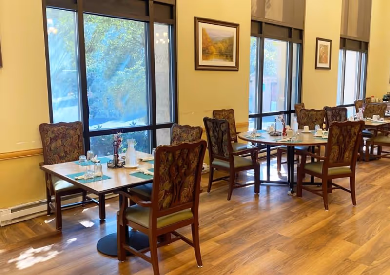 Sunlit dining room with multiple set tables and patterned chairs beside large windows.