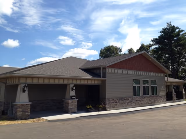 Exterior view of a single-story building with a sloped roof, stone and siding facade, several windows, and a covered entrance. The sky is partly cloudy and there are trees in the background.