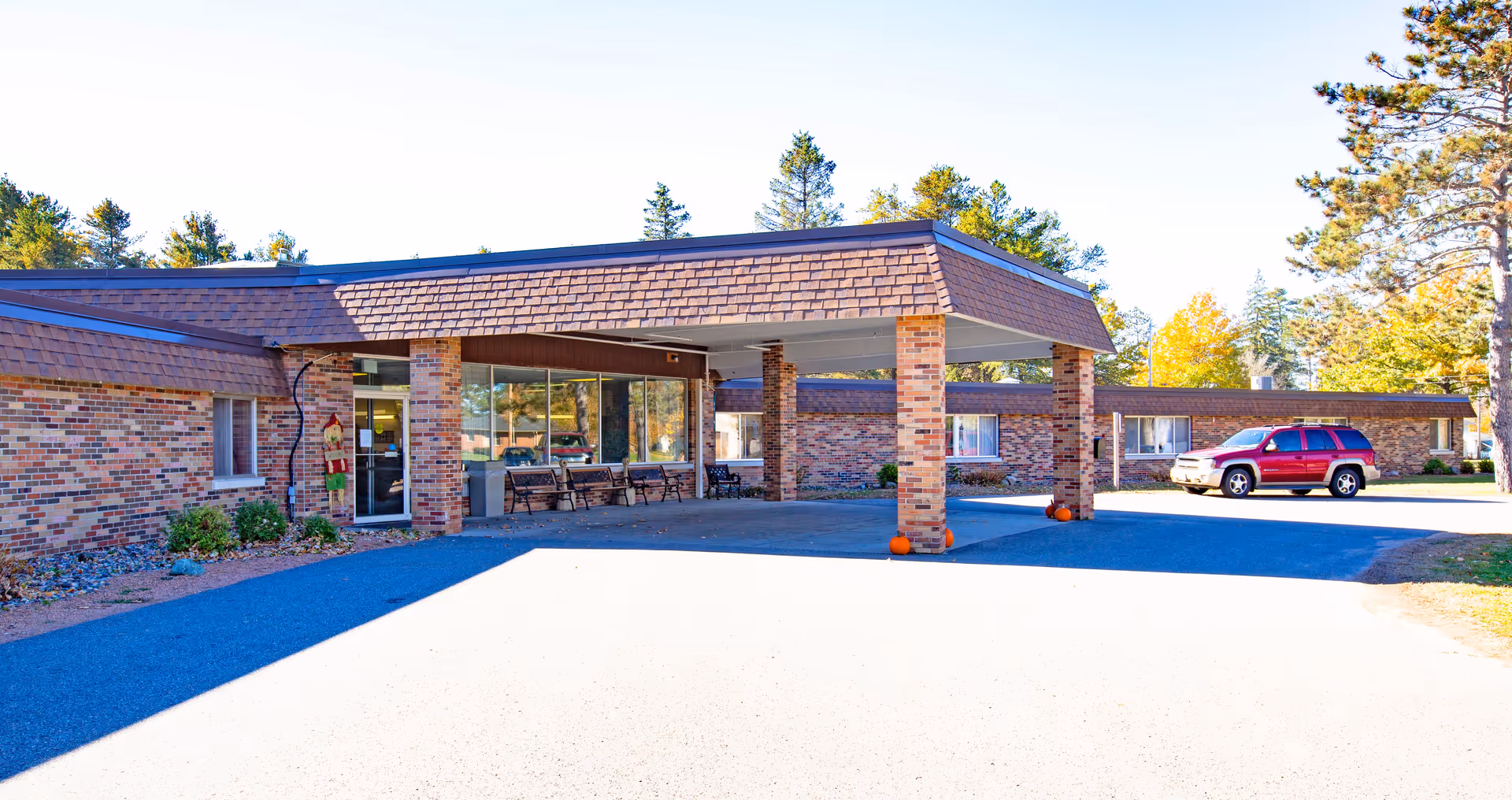 Exterior view of a single-story brick building with a covered entrance supported by brick pillars. There are benches near the entrance and a red SUV parked on the right side. Trees with autumn foliage are visible in the background under a clear blue sky.