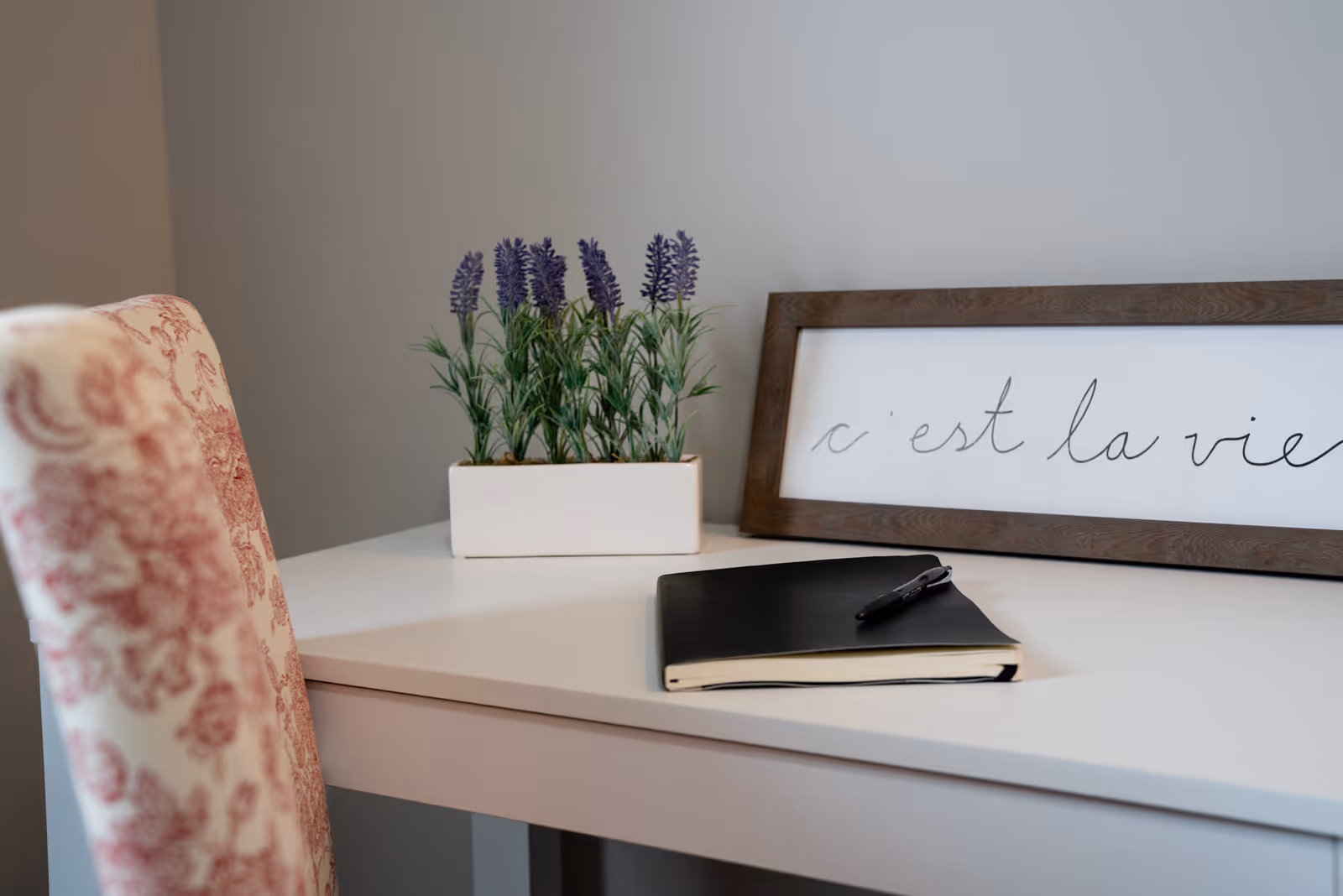 A white desk with a black notebook and pen on top, a rectangular white planter with purple flowers, and a framed sign with cursive writing that says 'c'est la vie'. A chair with a red and white floral pattern is partially visible on the left side.