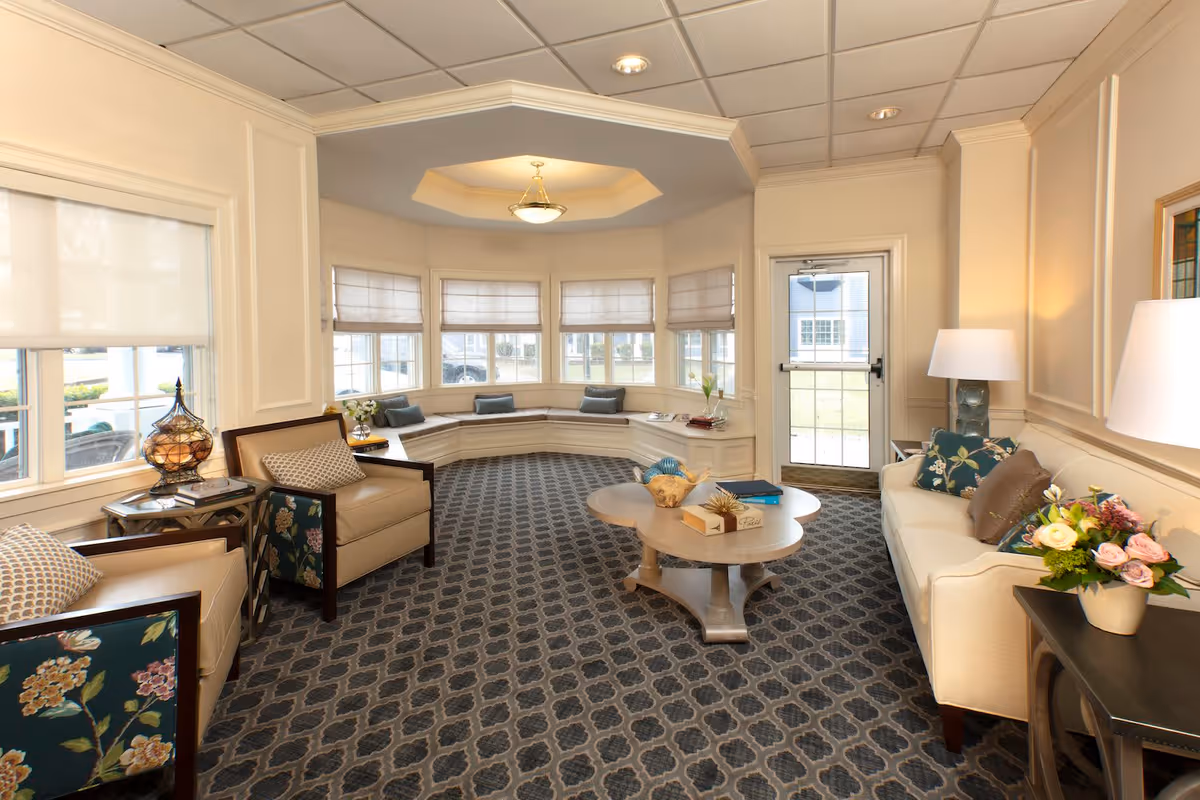 A cozy living room area in Heights Crossing Assisted Living featuring a patterned carpet, a white sofa with floral and brown cushions, two armchairs with floral upholstery on the sides, a round wooden coffee table with decorative items, a side table with a lamp and flowers, and a large bay window with cushioned seating underneath. The room is softly lit with ceiling lights and a table lamp, and there is a glass door leading outside.