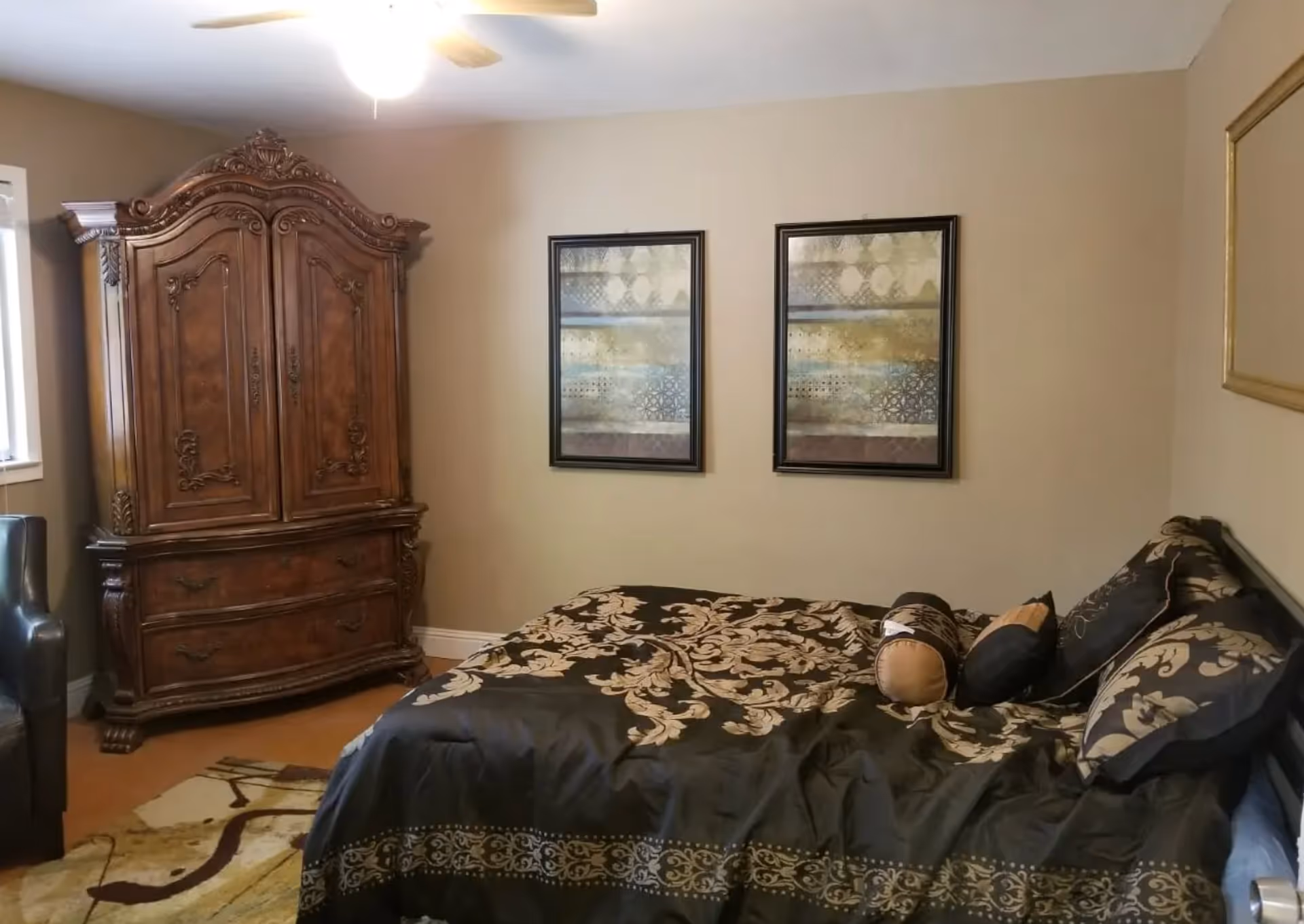 A bedroom with a decorative wooden armoire, patterned bedspread and pillows, two framed artworks on the wall, and a ceiling fan.