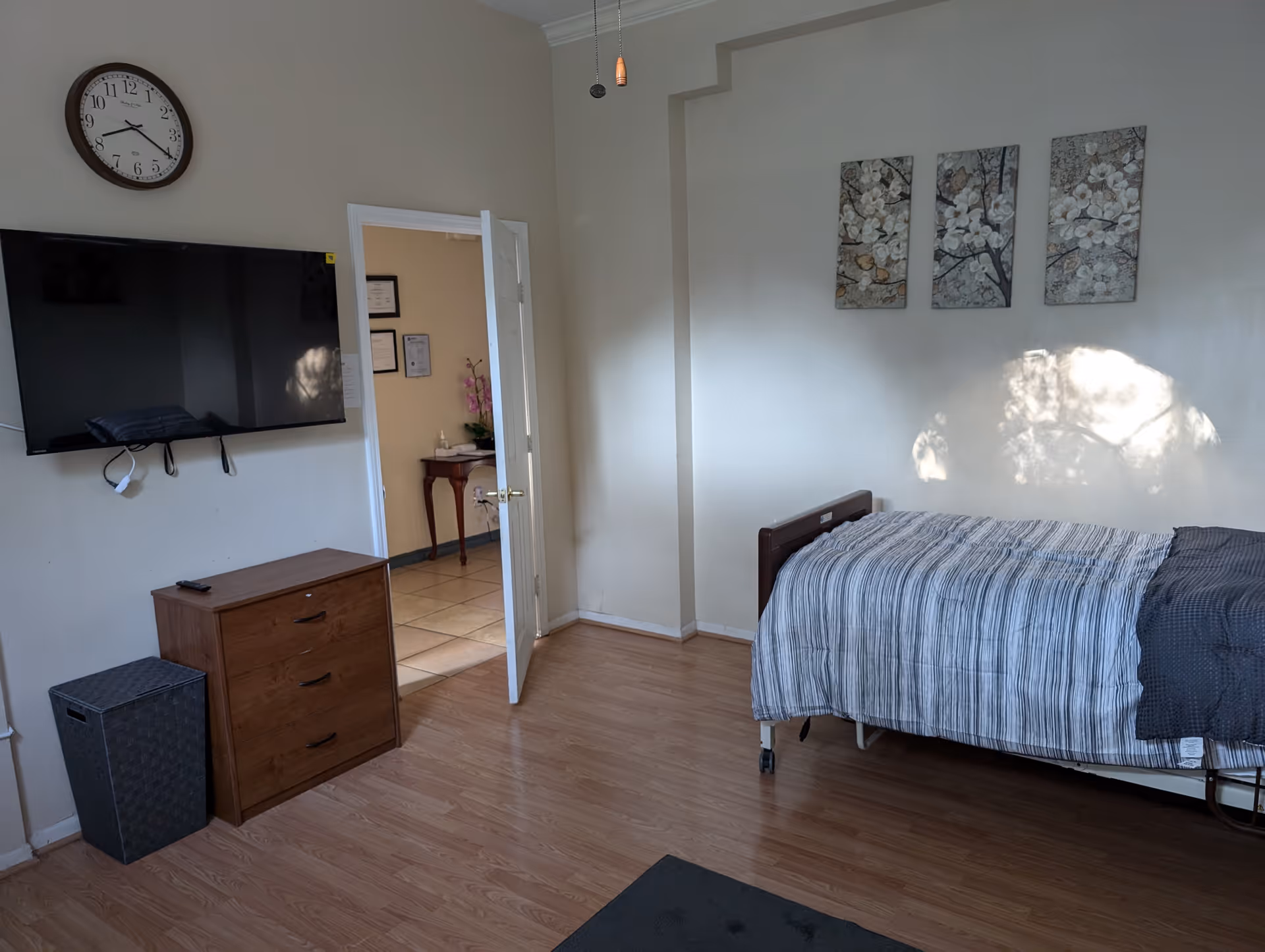 Furnished bedroom with a single bed, wall-mounted TV, dresser, clock, and an open door to a hallway.
