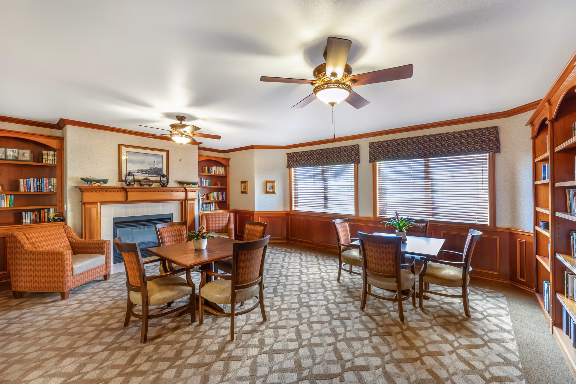 A cozy common area with two wooden tables each surrounded by four chairs, two armchairs, built-in wooden bookshelves, a fireplace with a mantel, and large windows with blinds and valances letting in natural light.
