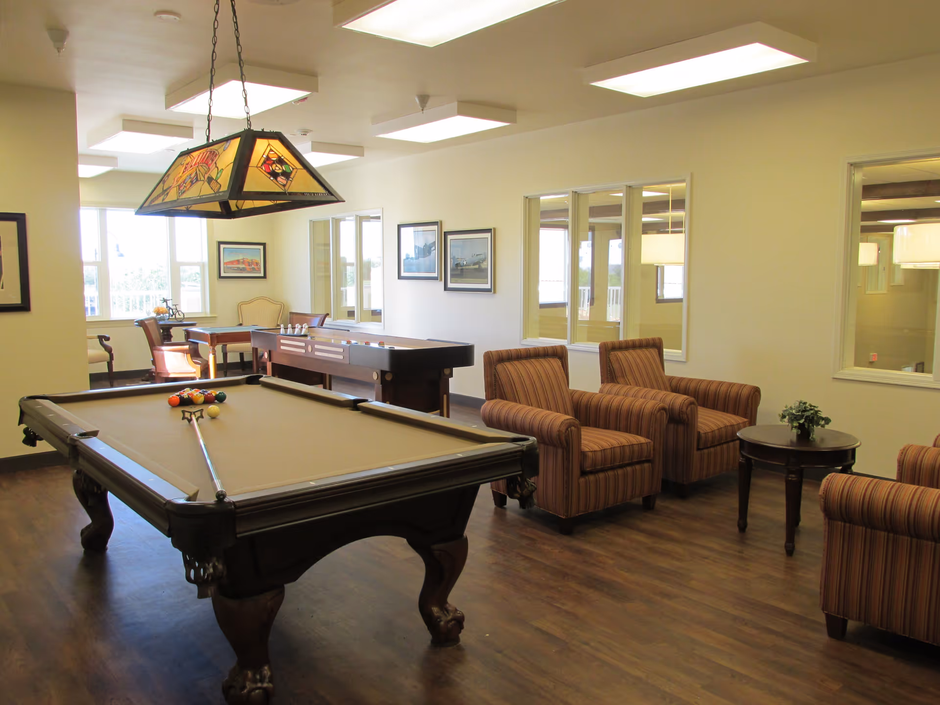 A recreational room with a pool table in the foreground, a shuffleboard table in the background, and several striped armchairs arranged around a small round table with a plant. The room has large windows, framed pictures on the walls, and a stained glass hanging light fixture above the pool table.