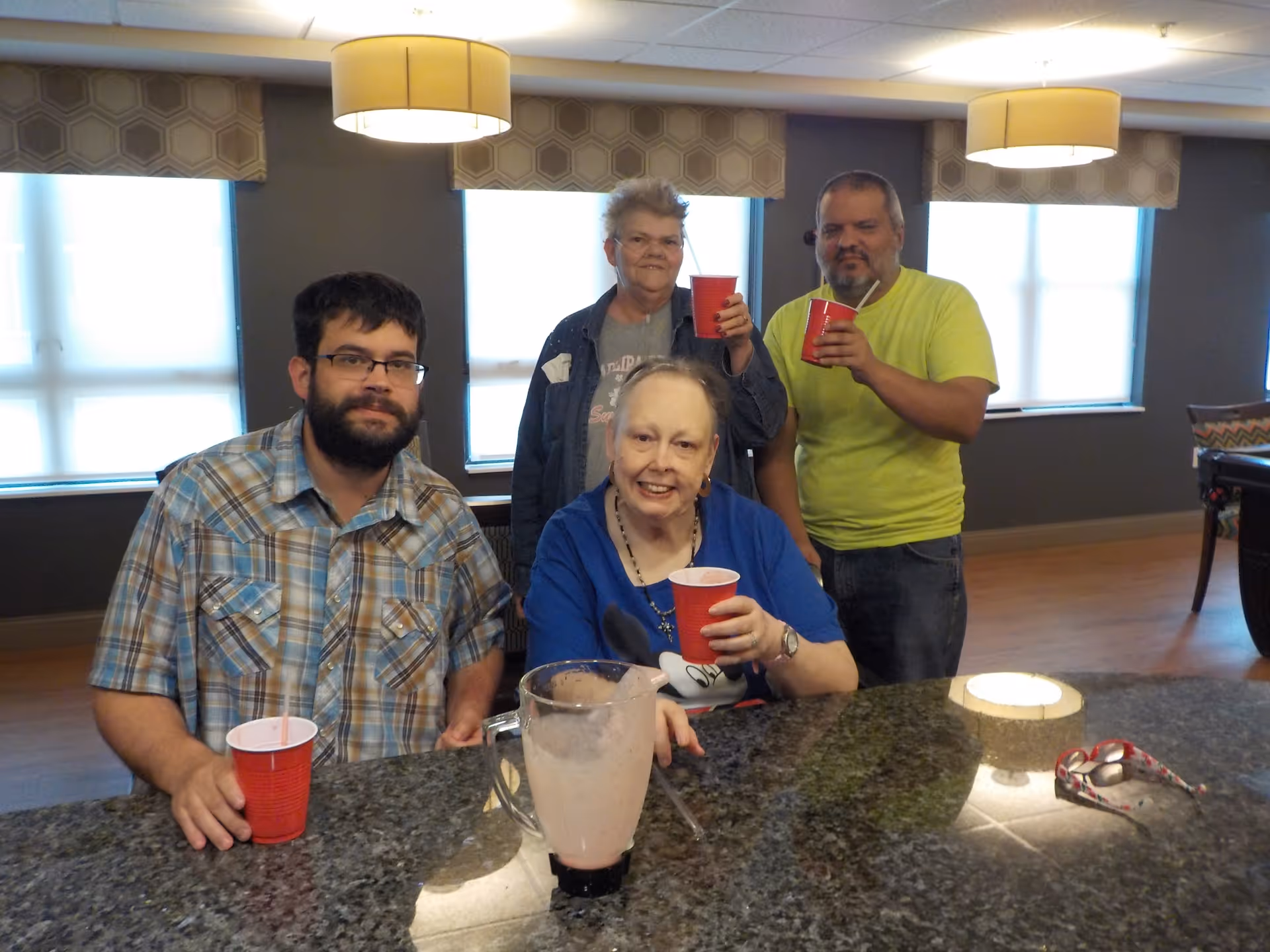 Four people gathered around a kitchen island in a well-lit room. Three of them are standing in the background holding red cups, while one person is sitting in the foreground also holding a red cup. There is a blender pitcher on the granite countertop and a pair of eyeglasses resting on the counter. The room has large windows with blinds and modern pendant lights hanging from the ceiling.