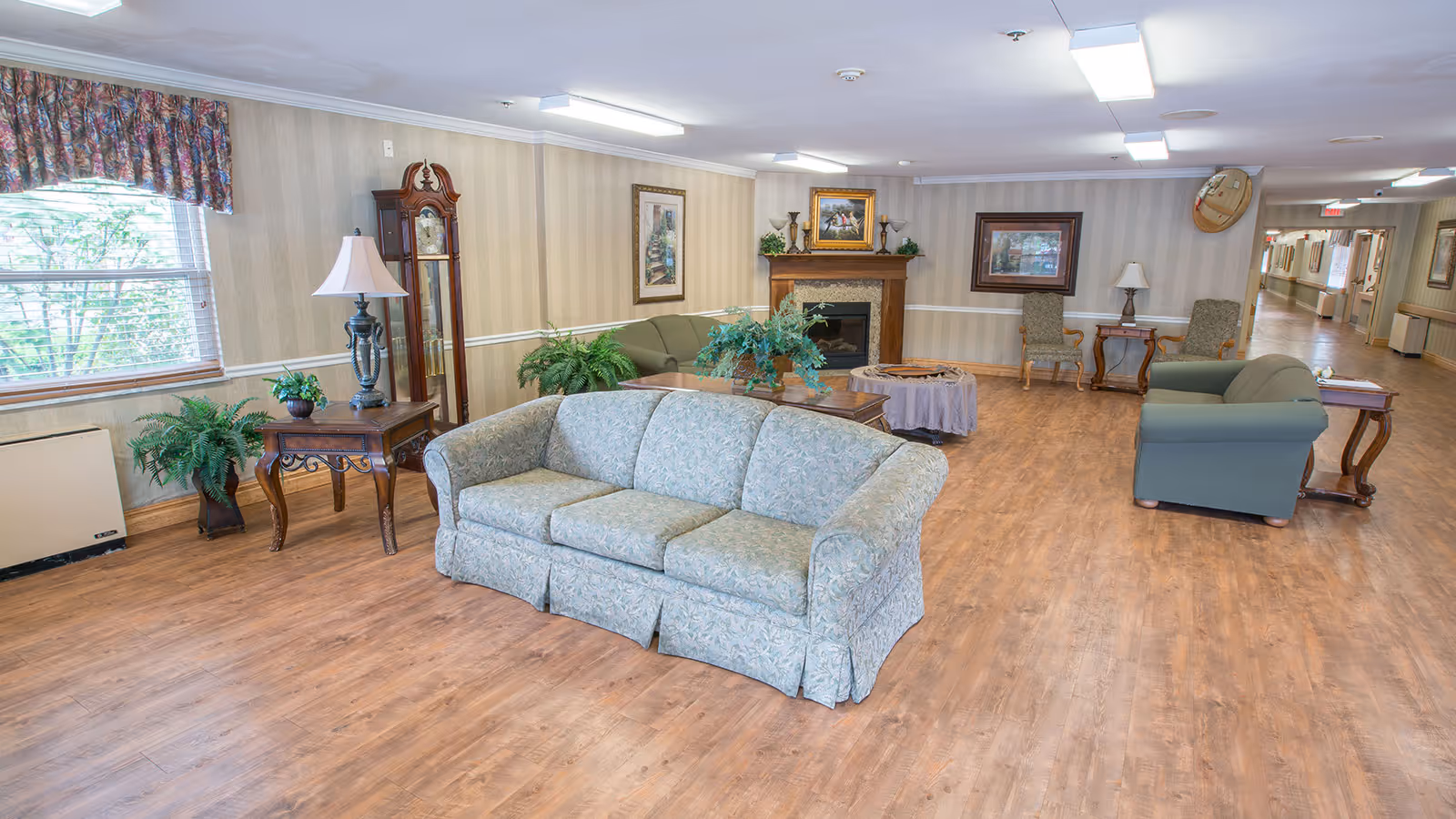 A spacious living room area in a nursing care center with wooden flooring, multiple sofas and armchairs, a grandfather clock, side tables with lamps, potted plants, framed artwork on the walls, and a fireplace. A long hallway is visible in the background.