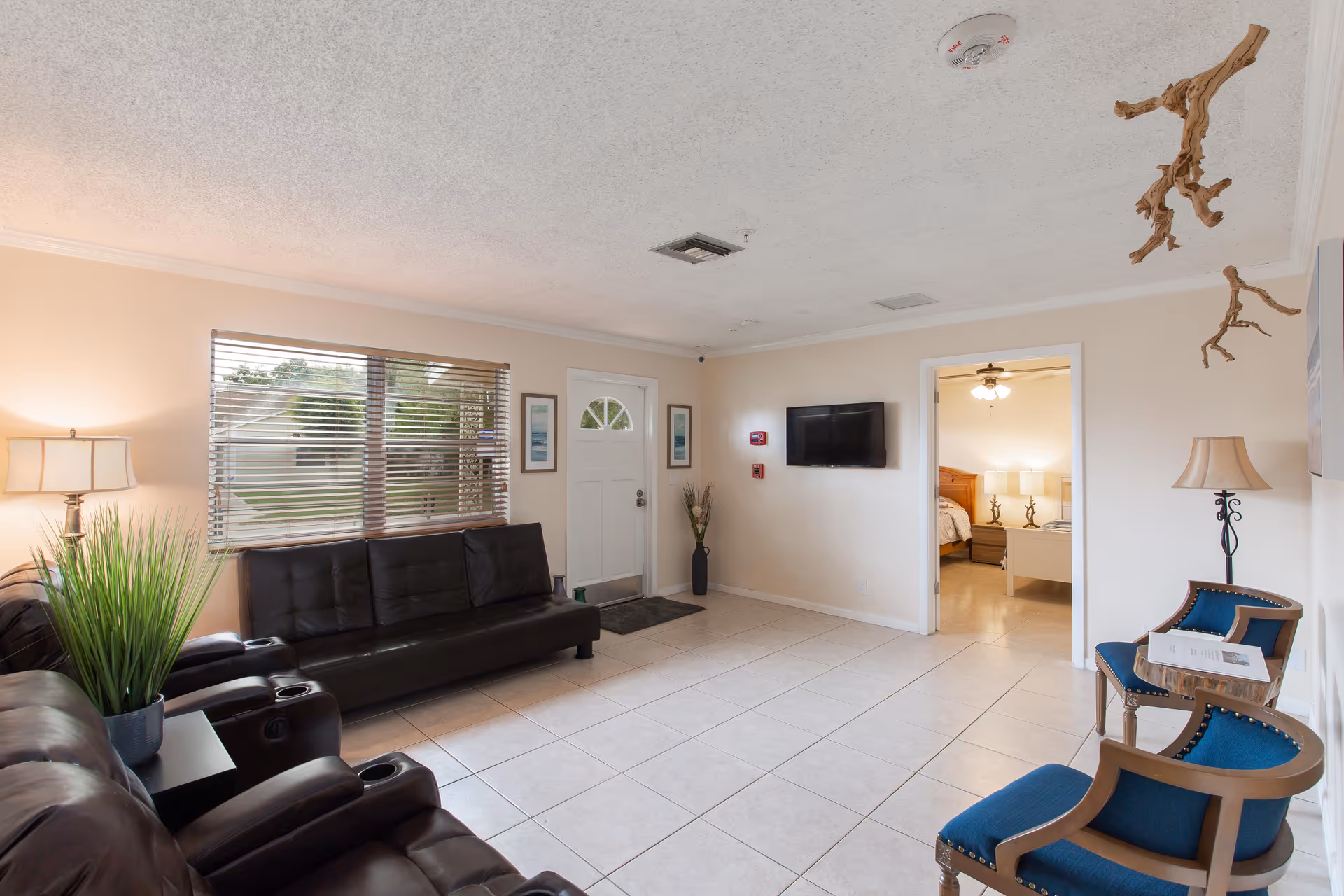 Bright common living room with leather sofas and blue armchairs, a wall-mounted TV, window blinds, and a doorway leading to a bedroom.