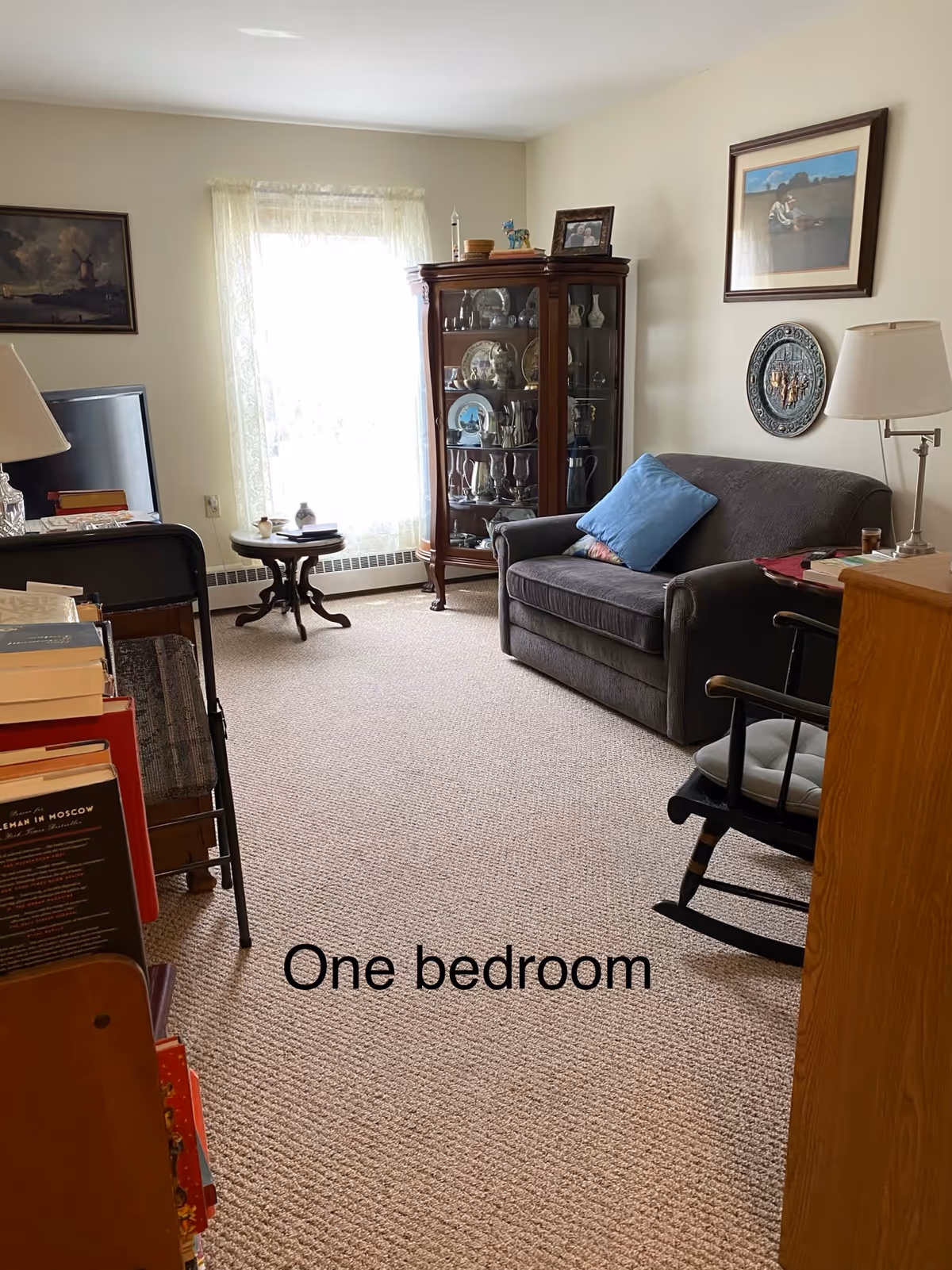 A furnished living area with a loveseat, rocking chair, curio cabinet and a window with lace curtain.
