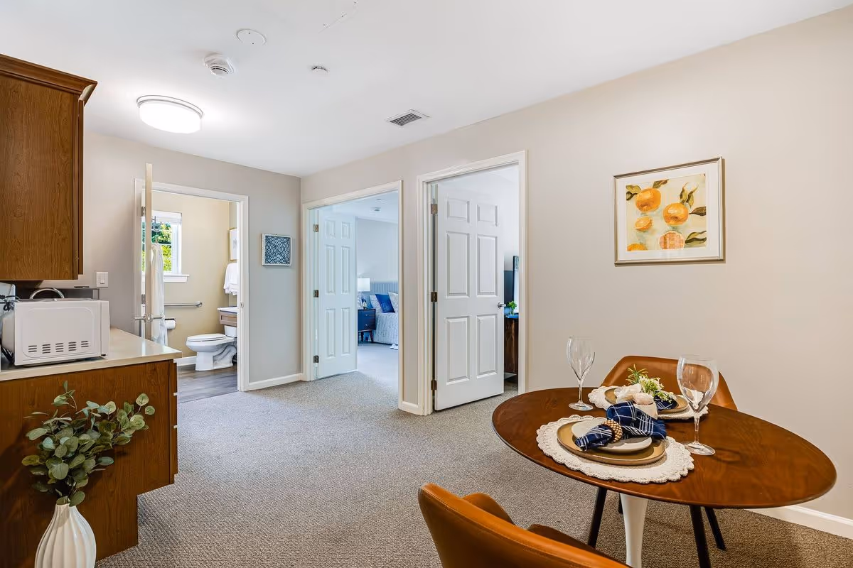 Interior view of a senior living facility showing a small dining area with a round wooden table set for two with plates, napkins, and glasses. To the left is a kitchenette with wooden cabinets and a microwave. In the background, there are two open doors leading to a bathroom and a bedroom. The walls are light-colored, and there is a framed artwork of oranges on the wall.