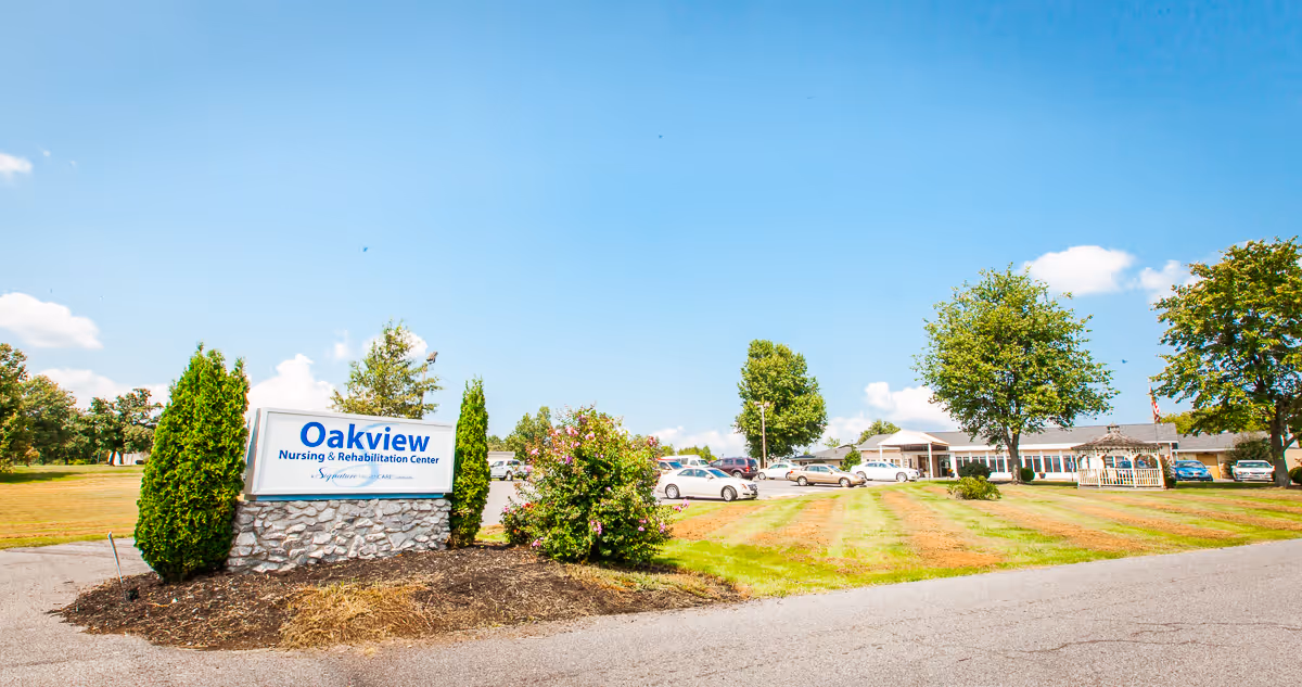 Exterior view of Oakview Nursing & Rehabilitation Center with its entrance sign, parking lot, lawn, and building under a blue sky.