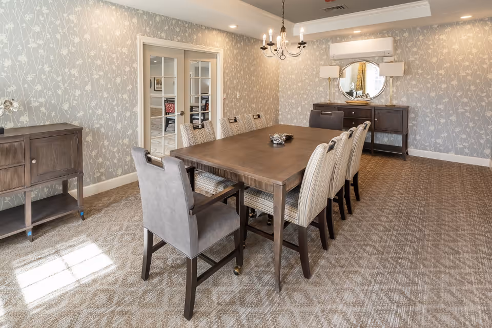 A dining room with a long wooden table surrounded by eight upholstered chairs. The room has patterned wallpaper with a floral design, a carpeted floor, a chandelier hanging above the table, and two wooden sideboards against the walls. There is a round mirror and two lamps on one sideboard, and double glass doors in the background.