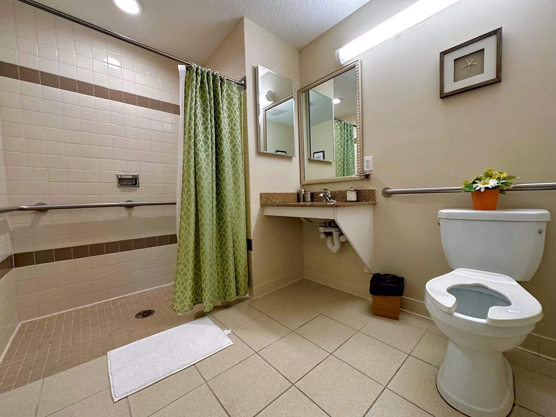 Accessible bathroom featuring a walk-in tiled shower with a green curtain, a sink and mirror, and a toilet with a raised seat and grab bar.
