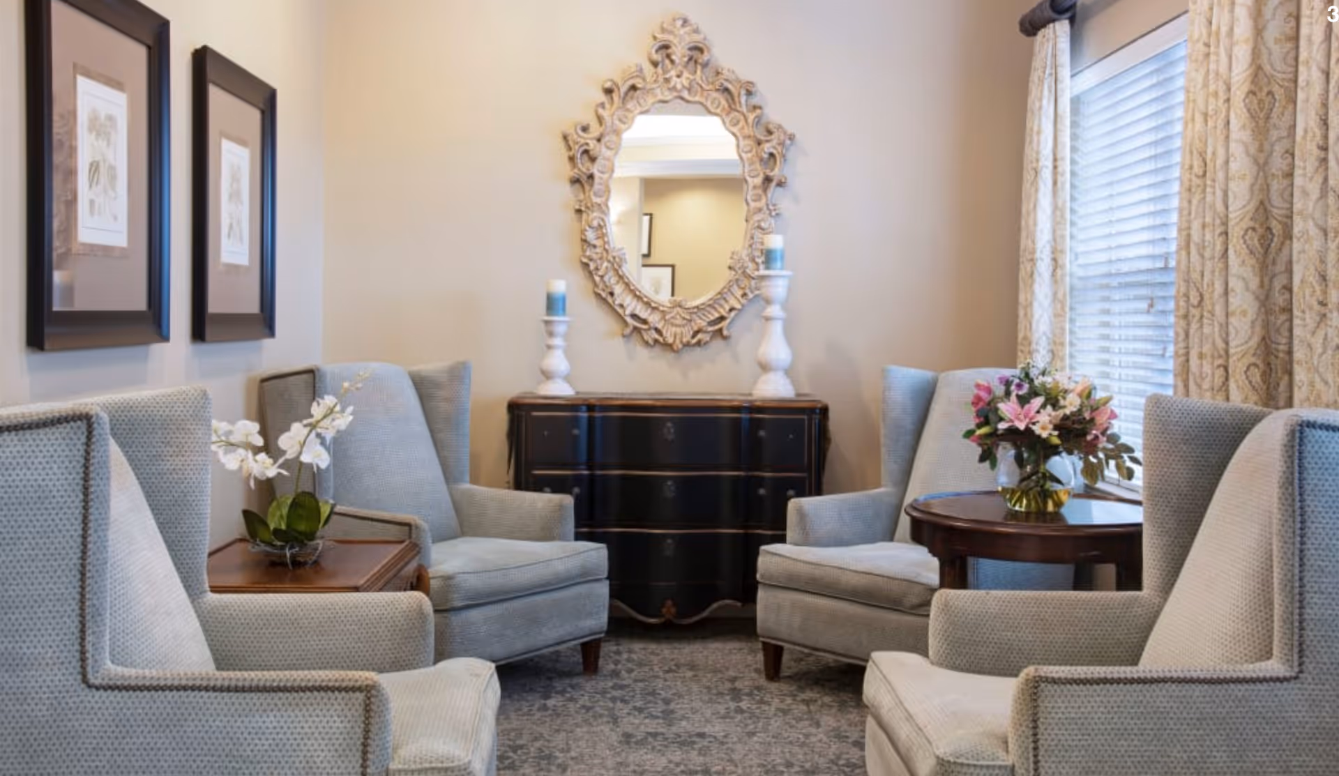 A cozy sitting area with four light blue upholstered armchairs arranged around two wooden side tables. One table holds a vase with white orchids, and the other has a bouquet of pink and white flowers. Behind the chairs is a black chest of drawers with two white candle holders and a decorative ornate mirror hanging above it. The room has beige walls, two framed botanical prints, and a window with patterned curtains letting in natural light.