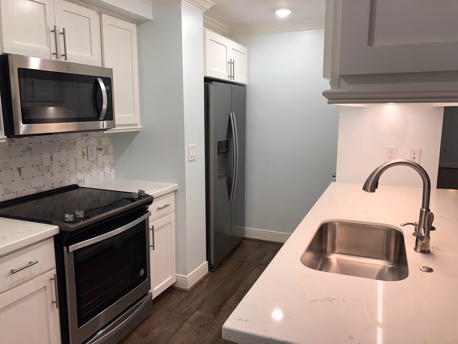 Modern kitchen with white cabinets, stainless steel microwave, stove, refrigerator, and a sink with a curved faucet on a white countertop. The floor is wooden and the walls are light-colored.