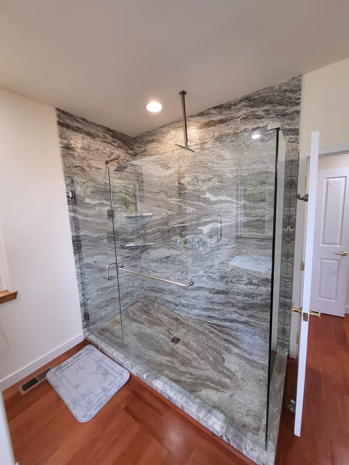 A modern bathroom shower with glass walls and a door, featuring a marble-patterned interior with built-in shelves and a metal grab bar. The shower has a ceiling-mounted showerhead and is adjacent to a wooden floor with a light gray bath mat outside the shower.