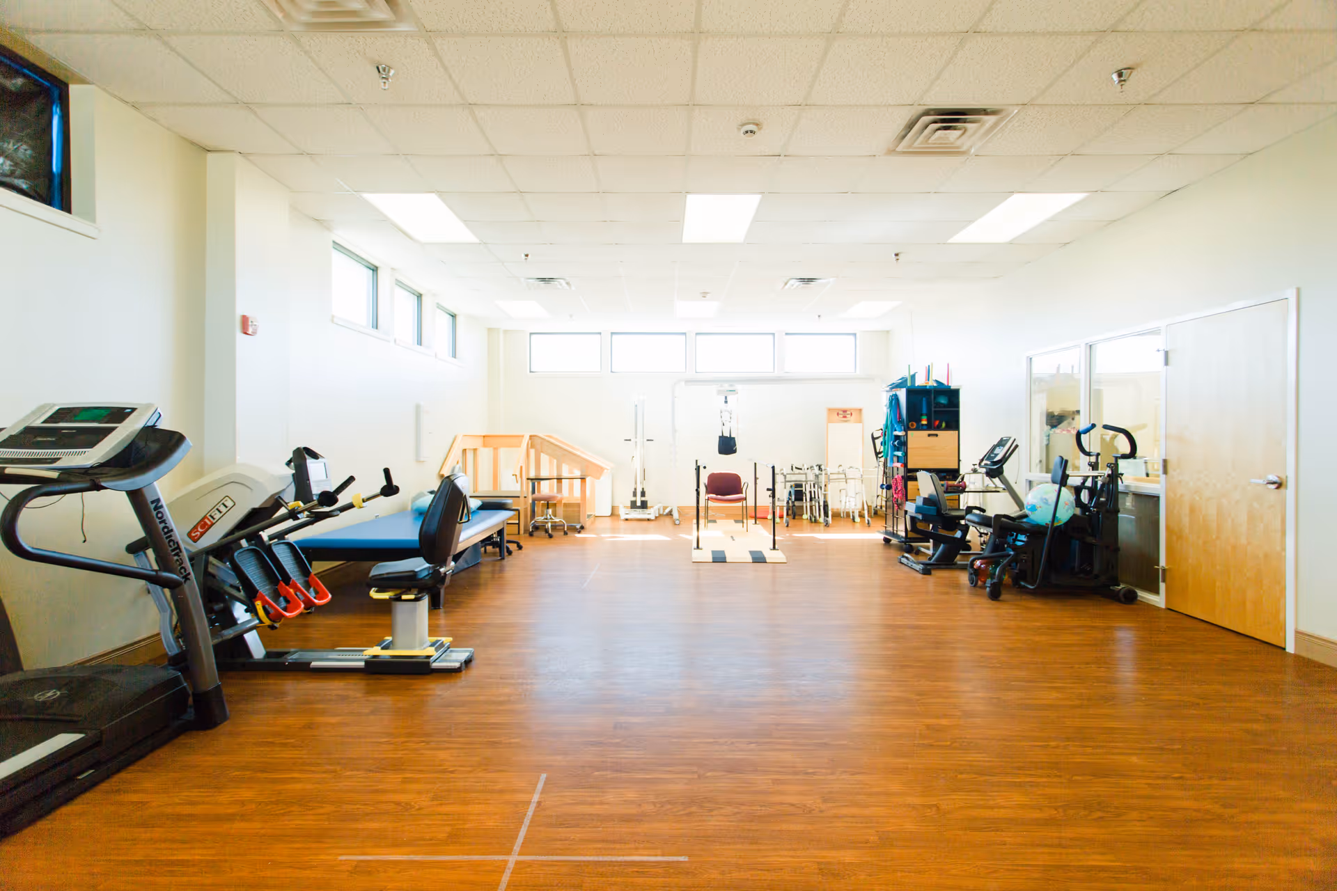 A spacious rehabilitation or physical therapy room with exercise equipment including a treadmill, stationary bikes, parallel bars, and other therapy tools. The room has wooden flooring, white walls, and large windows letting in natural light.