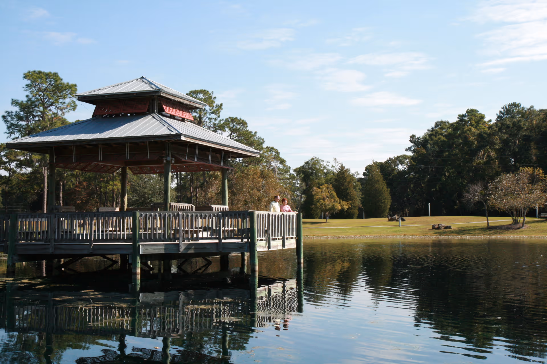 A wooden gazebo on a pier over a pond with two people standing on it and trees reflected in the water.