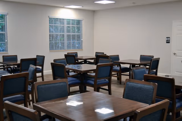 A dining room with multiple square wooden tables and blue cushioned chairs arranged neatly. The room has white walls, two windows with blinds, and a door with a restroom sign on it.