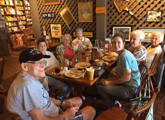 A group of elderly people and a younger woman sitting around a wooden dining table in a cozy restaurant setting. The table has plates with food remnants, drinks, and condiments. The background shows wooden lattice walls decorated with framed pictures and signs.