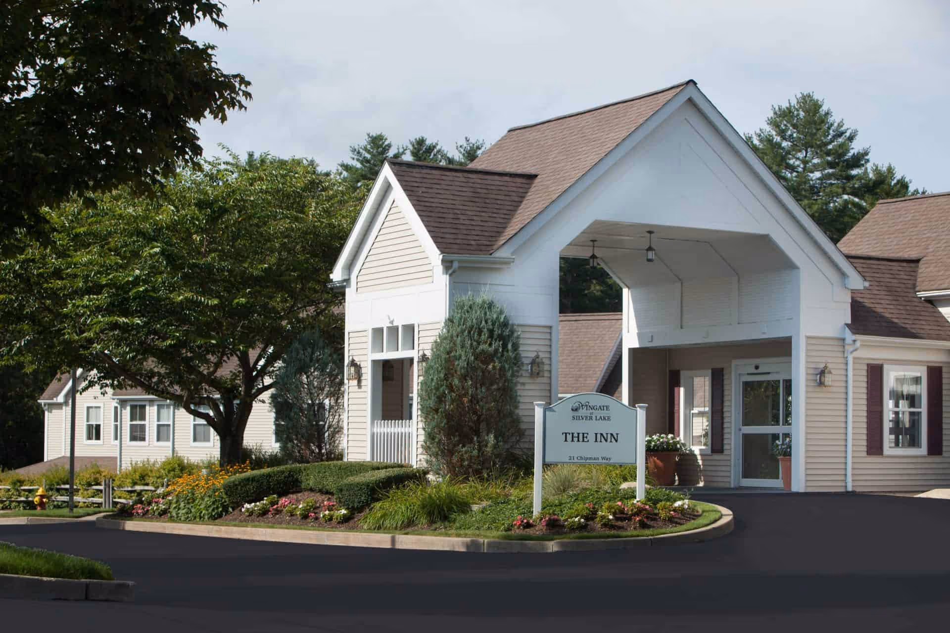 Exterior view of Wingate Residences at Silver Lake showing the entrance to The Inn with a covered driveway, surrounded by well-maintained landscaping including bushes, flowers, and trees.