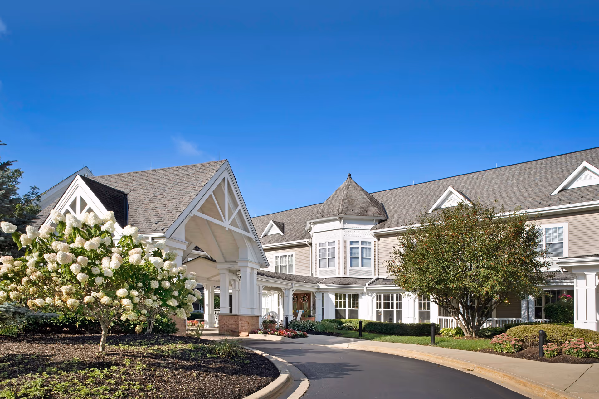 Exterior view of a senior living facility building with a covered entrance, surrounded by landscaped gardens with blooming white flowers and green trees under a clear blue sky.