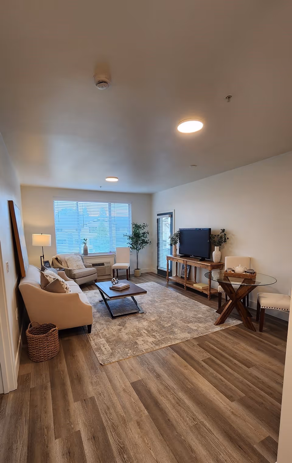A cozy living room with wooden flooring, a beige sofa, an armchair, a coffee table, a TV on a wooden console, a round glass dining table with two chairs, and large windows with blinds letting in natural light.