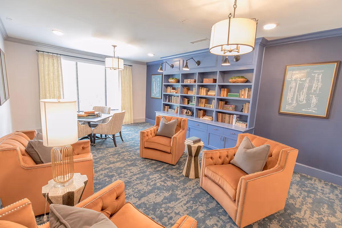 A cozy living room area with four orange upholstered armchairs arranged around a small side table with books. The room features a large blue built-in bookshelf filled with books and decorative items. There is a dining table with four chairs near a window with light-colored curtains. The walls are painted blue and white, and the room is well-lit with ceiling lights and a table lamp.