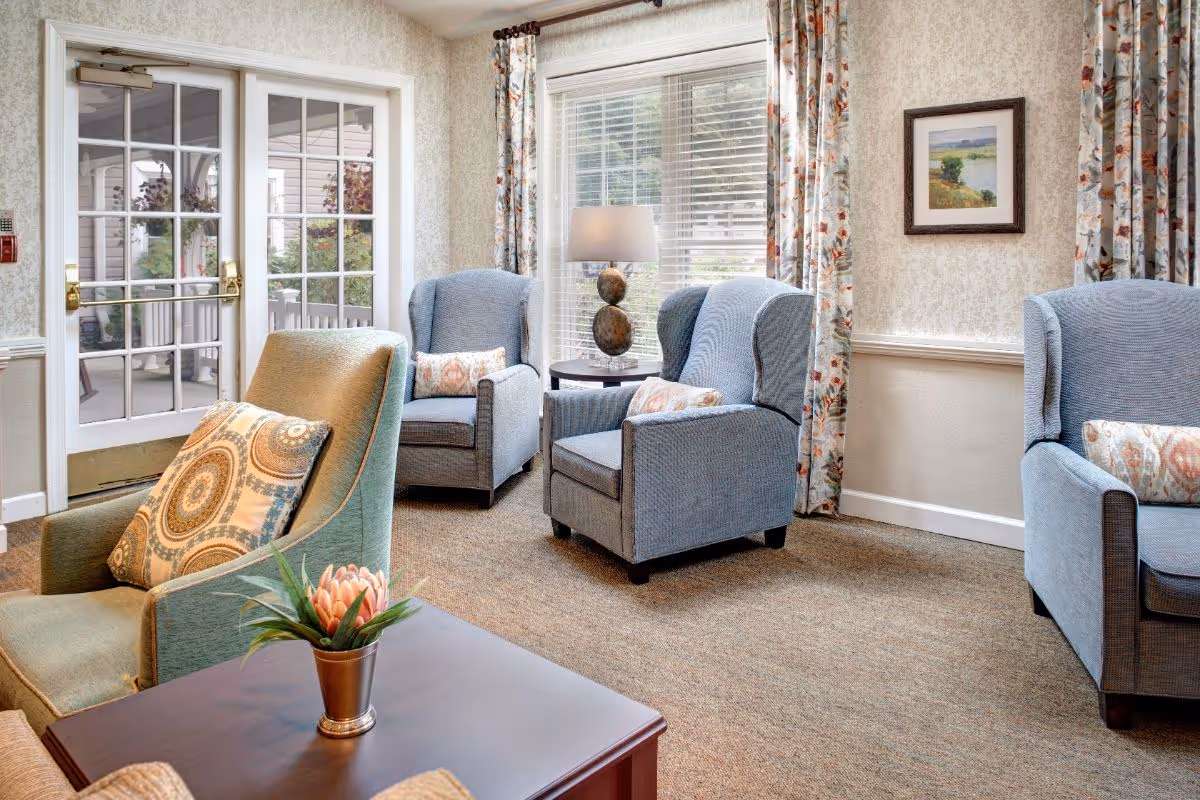 A bright lounge with upholstered armchairs, a side table with a lamp, floral curtains, and glass double doors.