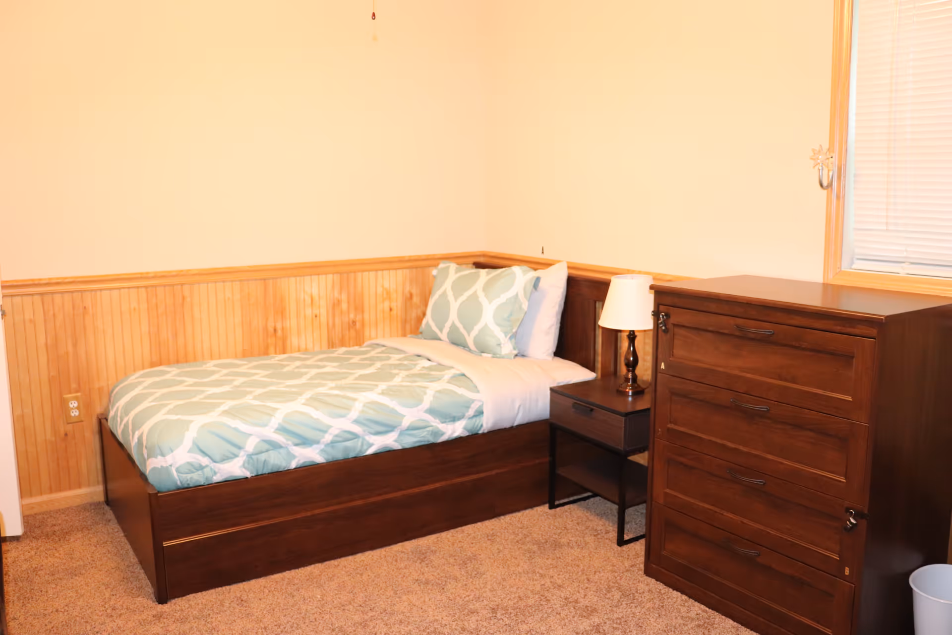 A small bedroom with a single bed featuring a green and white patterned comforter and pillows. Next to the bed is a dark wooden nightstand with a lamp on it. To the right of the nightstand is a matching dark wooden dresser with multiple drawers. The room has beige walls with wooden wainscoting and a window with closed blinds.