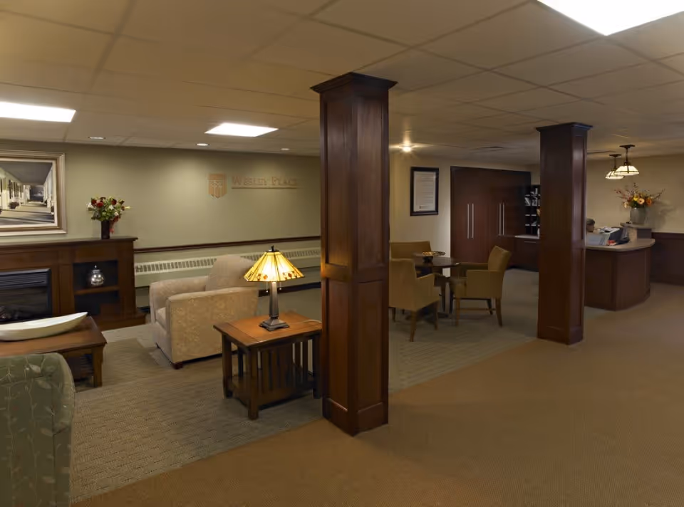 A cozy and warmly lit common area in a senior living facility featuring comfortable armchairs, a wooden coffee table with a lamp, a fireplace, and a reception desk in the background. The space has wooden pillars, carpeted floors, and decorative flowers on a side table and near the reception.