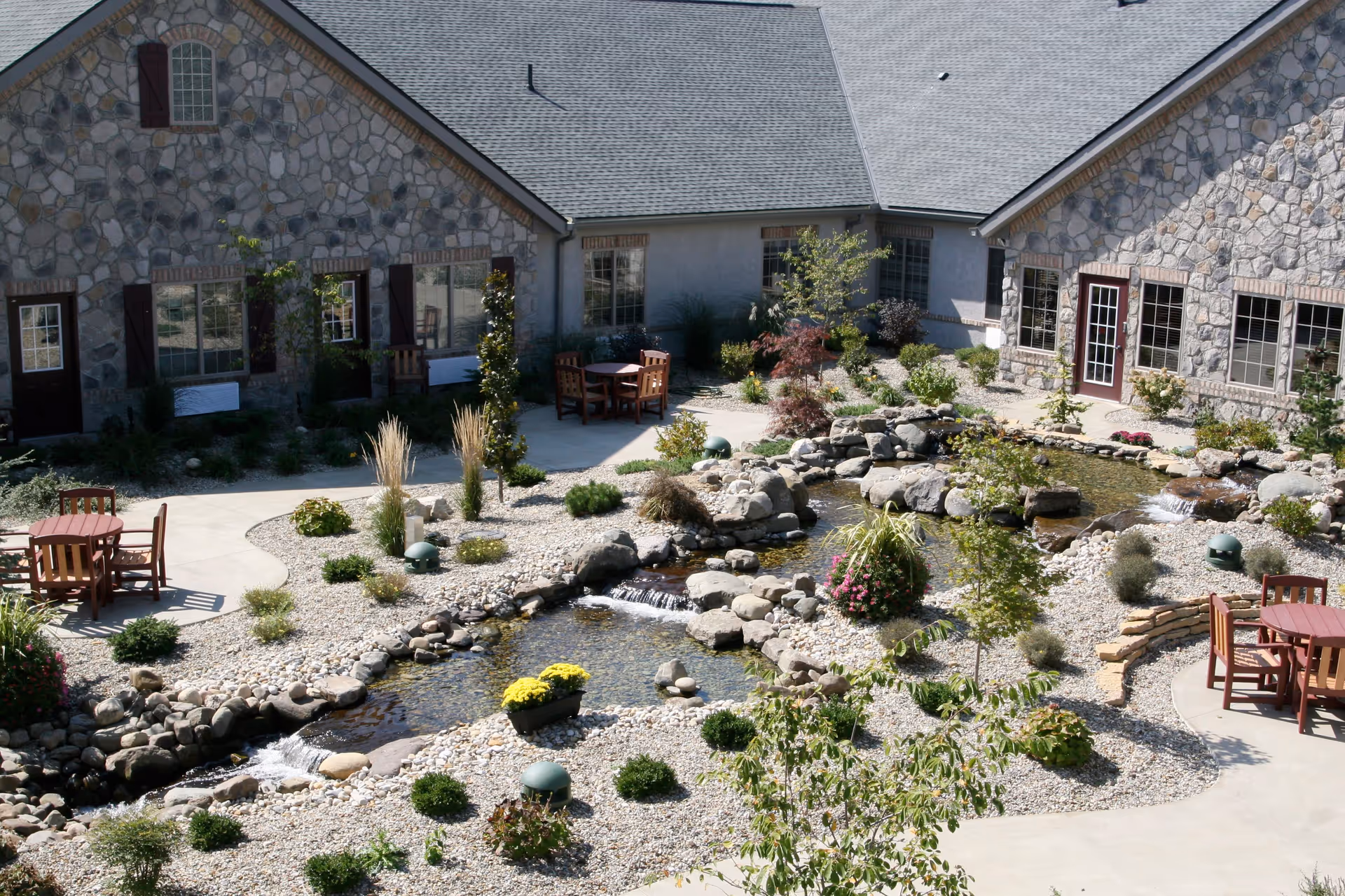 Outdoor courtyard area at Inn at Bear Trail featuring a landscaped garden with a small stream and rock formations. There are several wooden tables and chairs placed on concrete patios surrounded by various plants and shrubs. The building exterior is made of stone with multiple windows and doors.