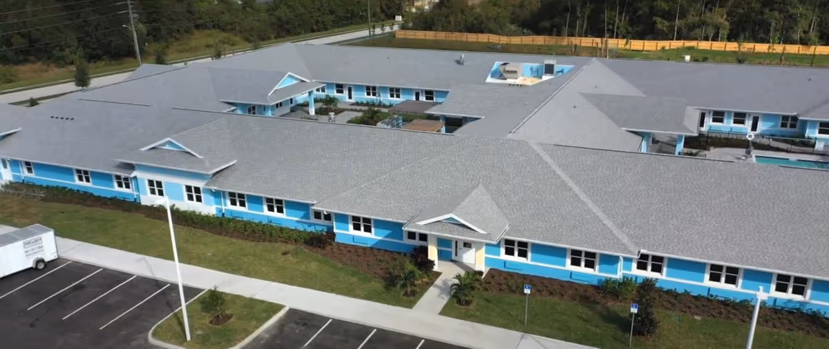 Aerial view of a single-story assisted living facility with a gray roof and bright blue exterior walls. The building is surrounded by a parking lot with marked spaces and landscaped greenery. The facility has multiple wings and a central courtyard area.