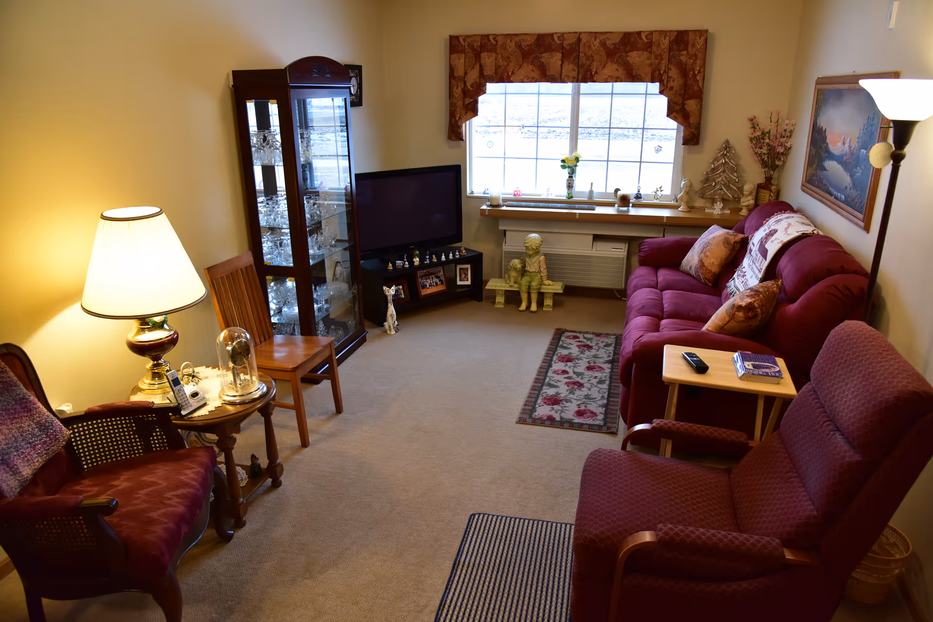 A cozy living room with a red sofa, two armchairs, a wooden chair, a glass display cabinet filled with glassware, a TV on a stand, a window with a decorative valance, and various decorative items including a lamp, figurines, and framed pictures.