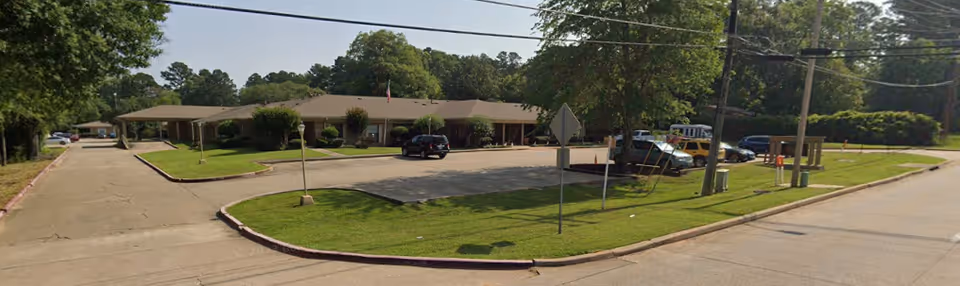 Exterior view of a single-story building with a parking lot in front, surrounded by trees and greenery under a clear sky.
