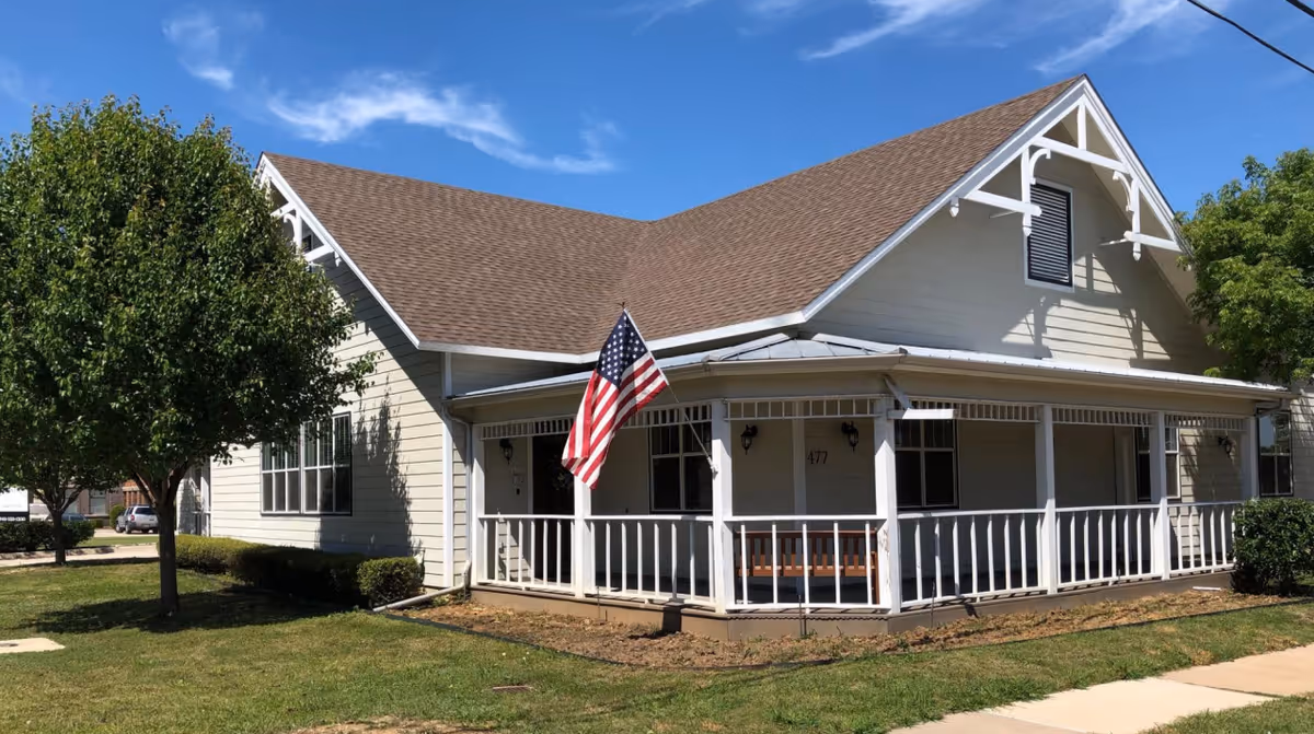 Front exterior of a pale yellow house-like building with a covered porch, an American flag, and surrounding lawn under a blue sky.