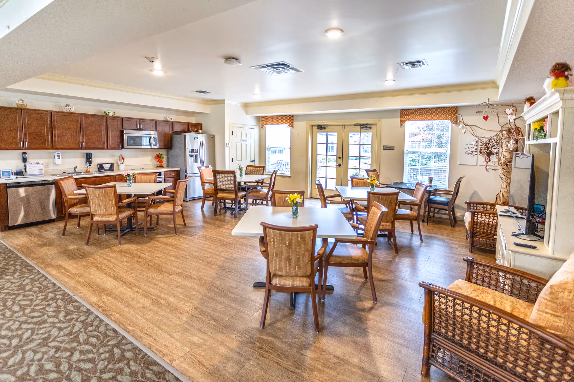 A bright and spacious dining area in a senior living facility with several square tables and wooden chairs arranged neatly. The room features a kitchen area with wooden cabinets, a microwave, a refrigerator, and a dishwasher. There are large windows and glass doors letting in natural light, and the floor is a combination of wood and carpet. The room is decorated with small flower vases on the tables and a decorative tree with hanging ornaments near a television and seating area.